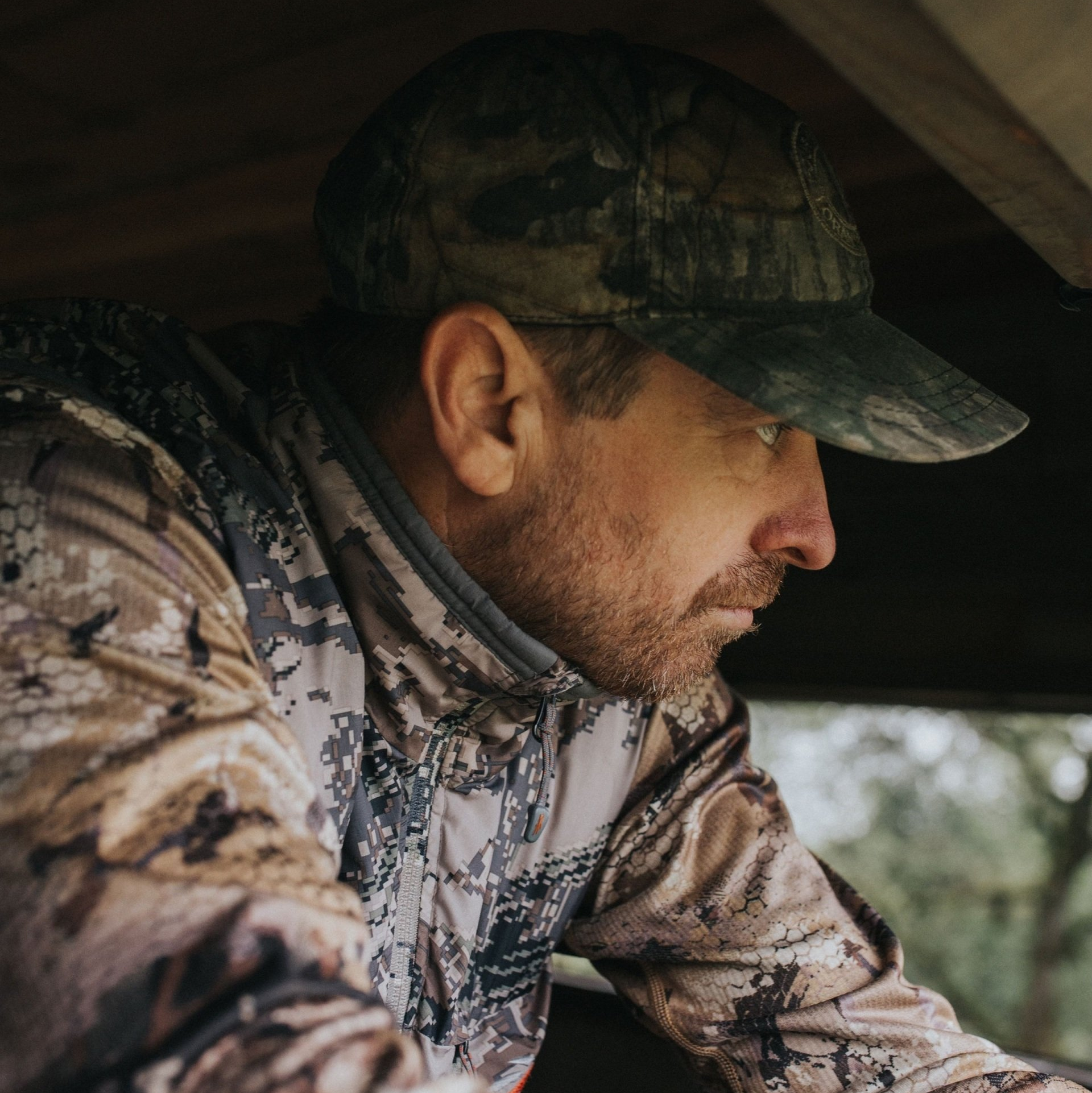 A man wearing a camo shirt and hat is looking out of a blind.