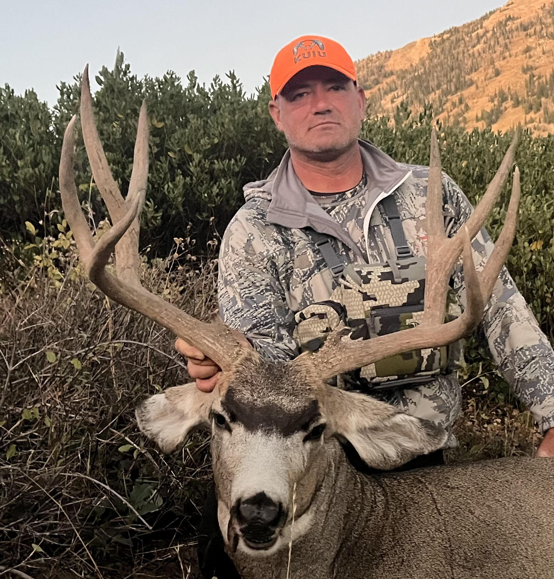 Man in camo holds large buck with impressive antlers in a mountainous setting.