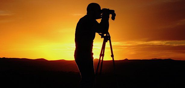 Silhouette of a person with a camera on a tripod at sunset. Orange sky and dark landscape.