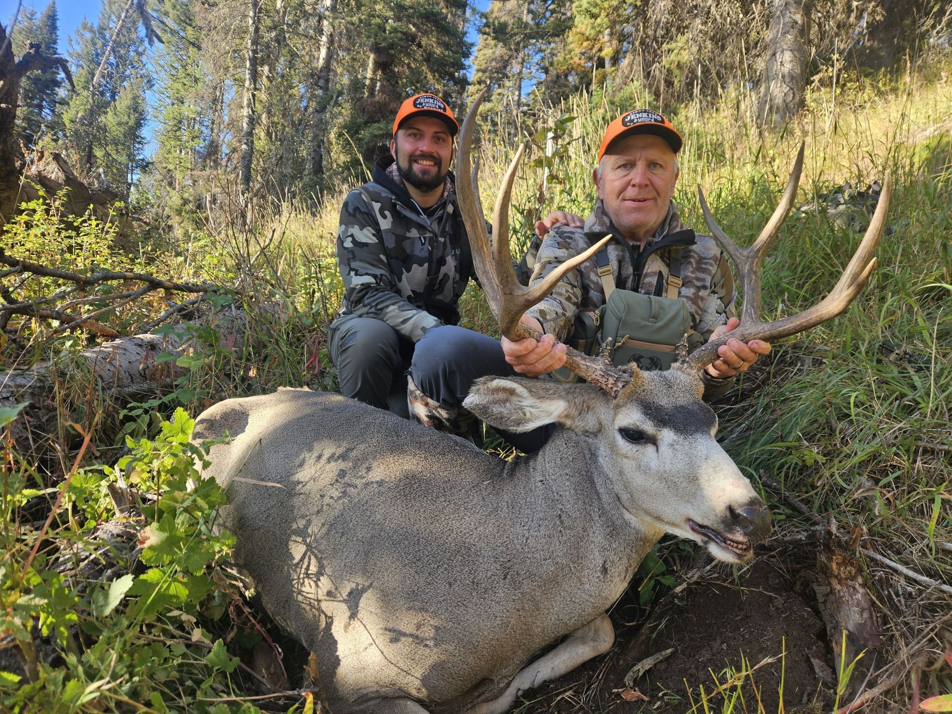 Two men pose with a large buck deer in a forest setting. The men are wearing camo and smiling.
