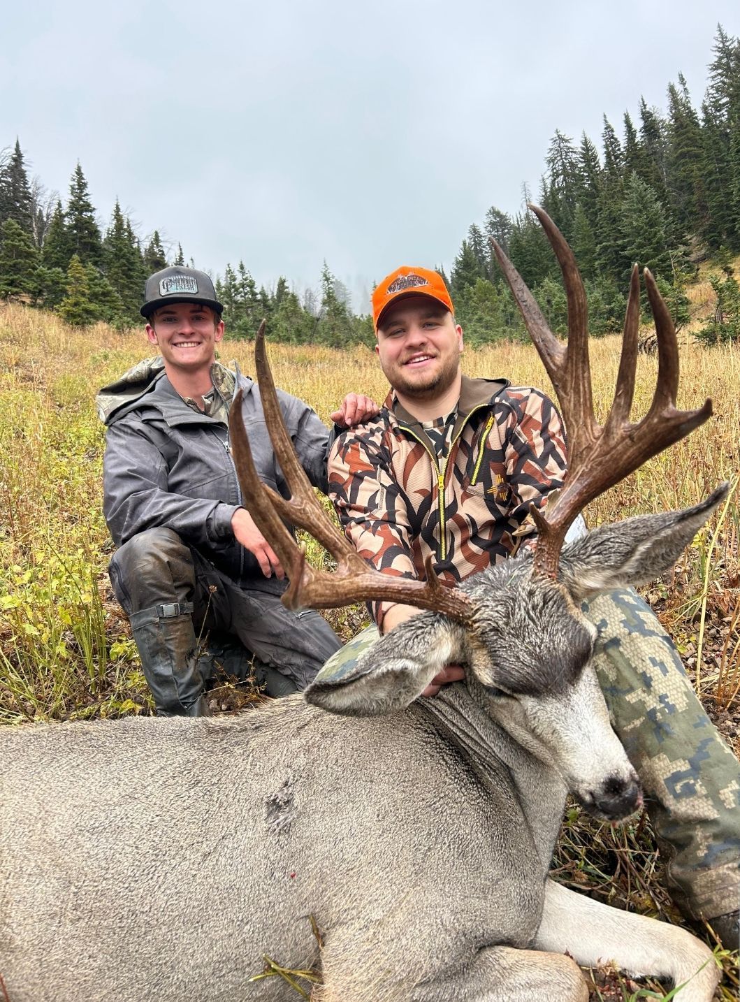 Two hunters pose with a large buck. One kneels in gray rain gear; the other smiles in camo and orange. Setting is a grassy hill.