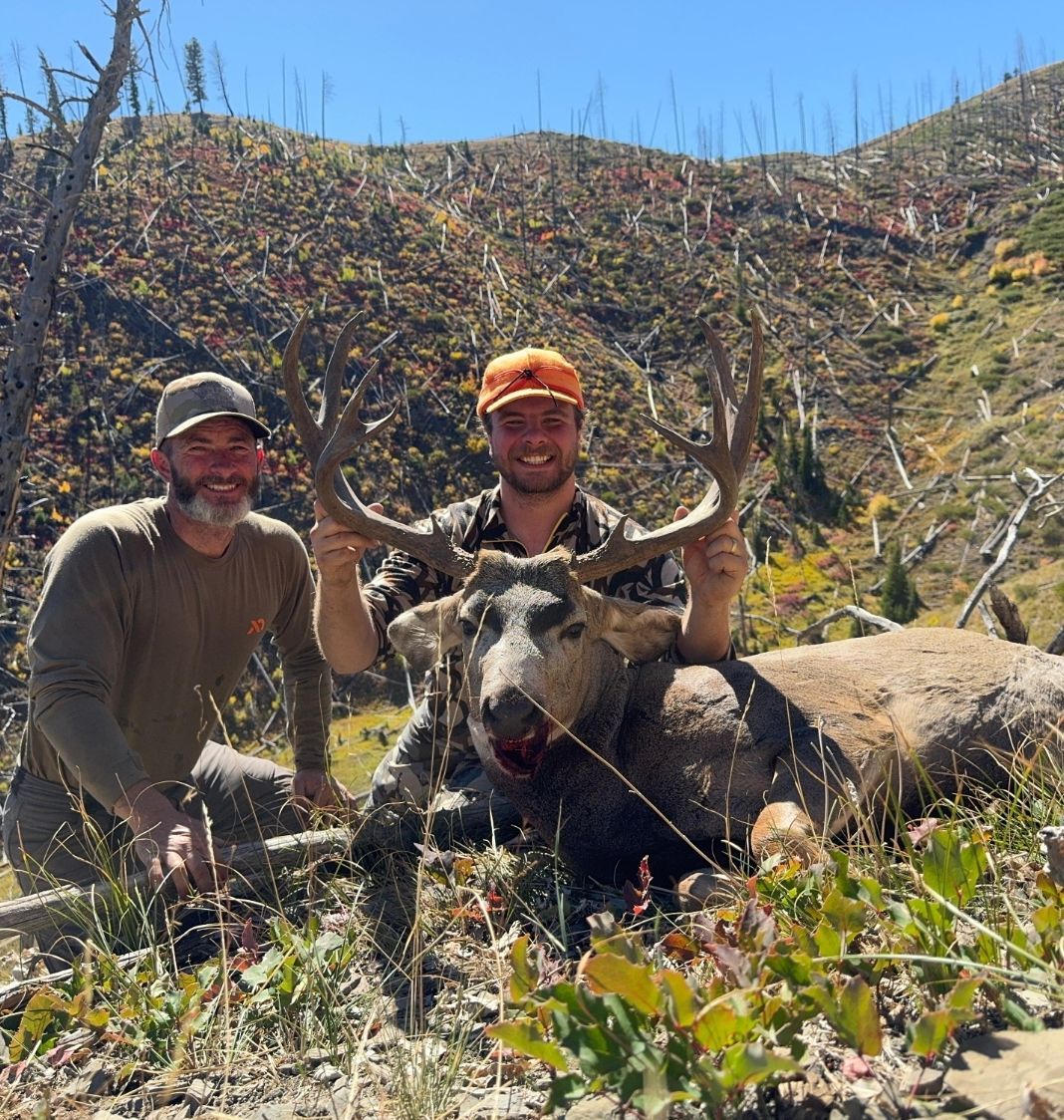 Two men with a large buck, antlers prominent. Mountain setting with dry vegetation. One man kneels, other holds the deer.