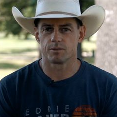A man wearing a cowboy hat and a blue shirt is looking at the camera.