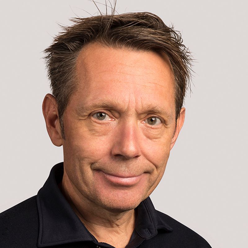 Man with short, light brown hair smiles, wearing a dark blue shirt, against a gray background.