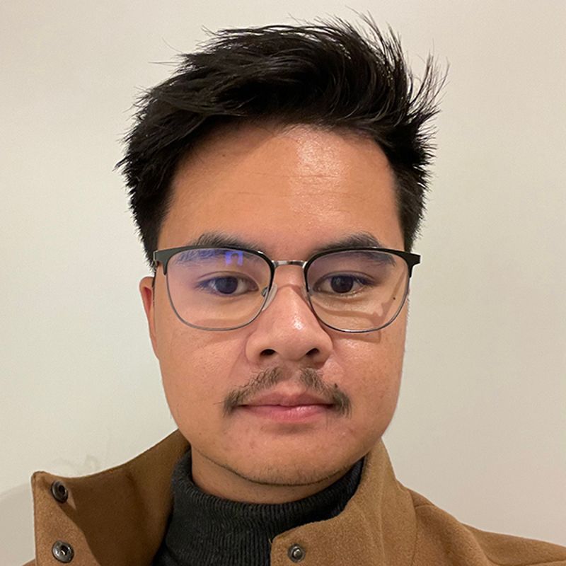 Man with glasses and mustache looking at the camera, wearing a brown jacket and black turtleneck.