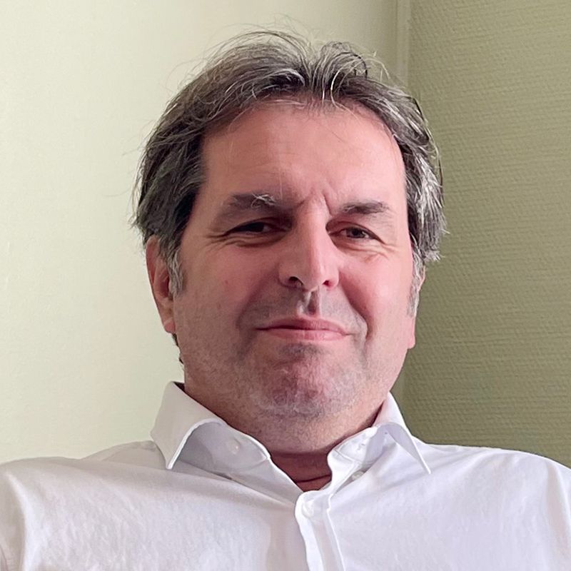 A man in a white shirt smiles at the camera. He has graying hair and is indoors.