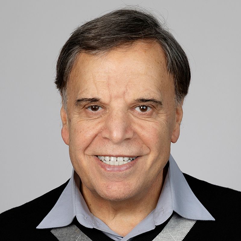 Smiling older man with dark hair, wearing a black sweater over a light blue collared shirt.