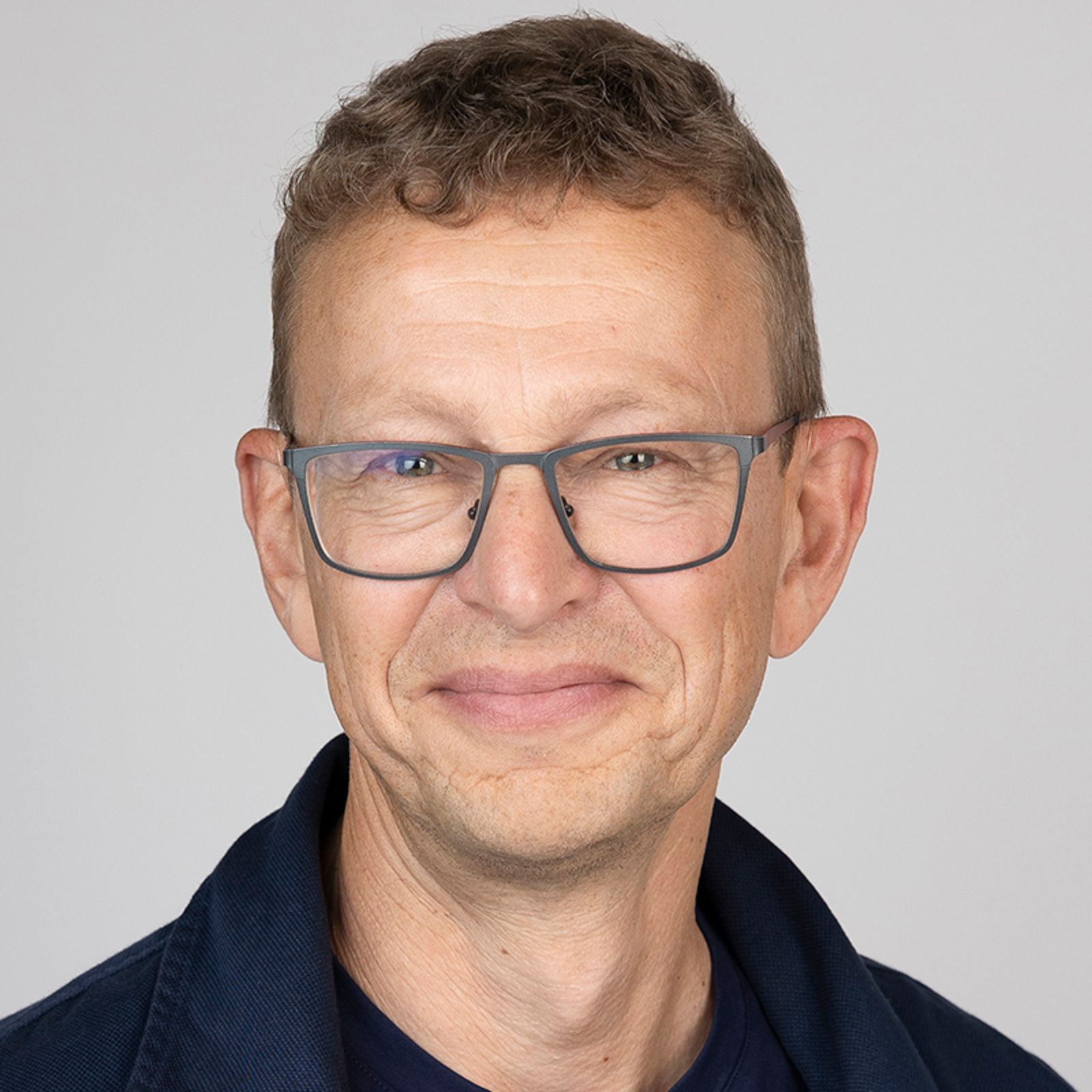Man with glasses, smiling, wearing a dark blue jacket, against a white background.