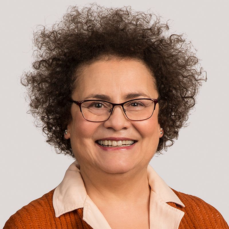 Woman with curly brown hair and glasses smiling, wearing an orange sweater and a white shirt.