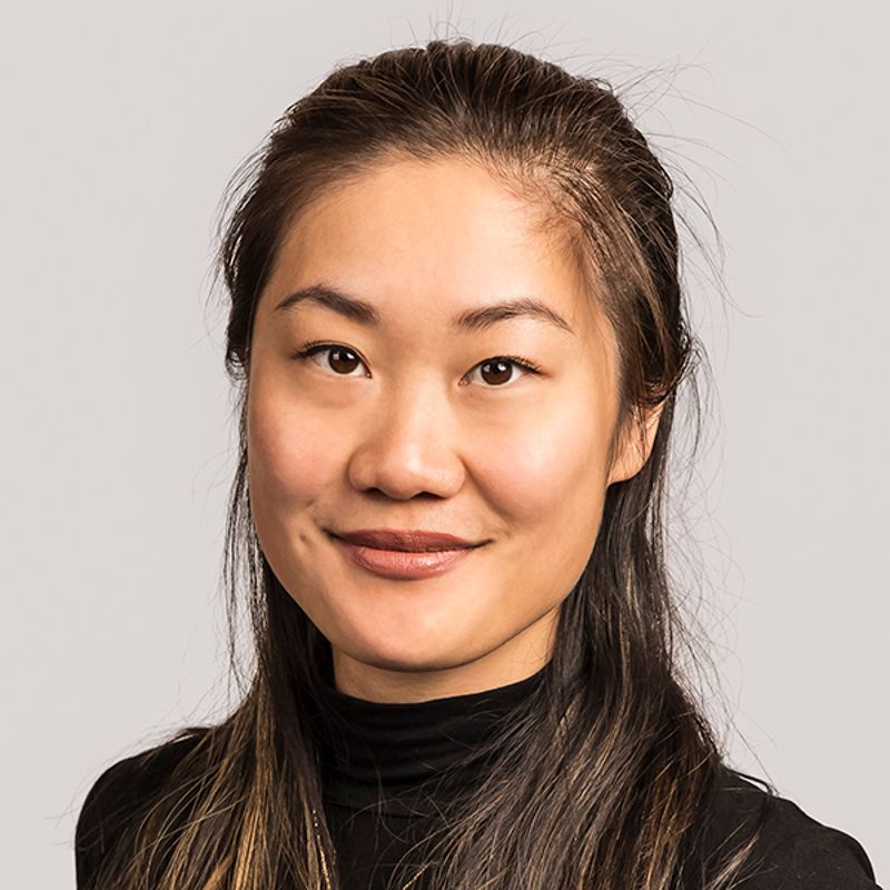 Woman with long dark hair smiles, wearing a black turtleneck, set against a gray background.