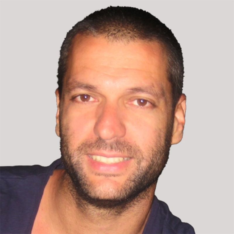 A man with short brown hair and a beard smiles. He wears a dark blue shirt against a gray background.