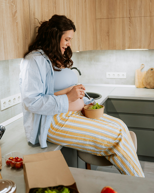 Person sitting on a kitchen counter, unpacking groceries near a bowl of vegetables.