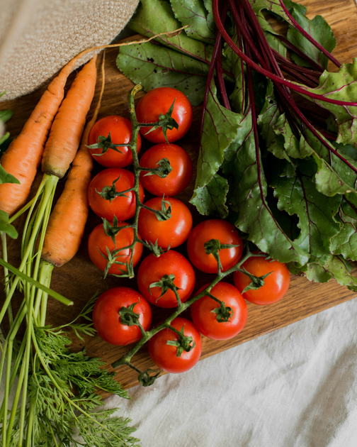 Fresh carrots, cherry tomatoes, dill, and beet greens arranged on a wooden cutting board.