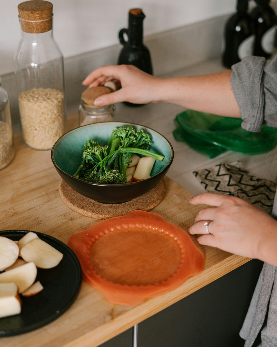 Hand placing broccoli in a bowl on a kitchen counter with plates and bread nearby