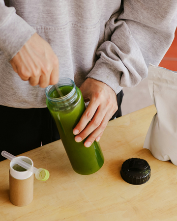 Hands pouring green smoothie into a bottle on a wooden table with blender parts nearby