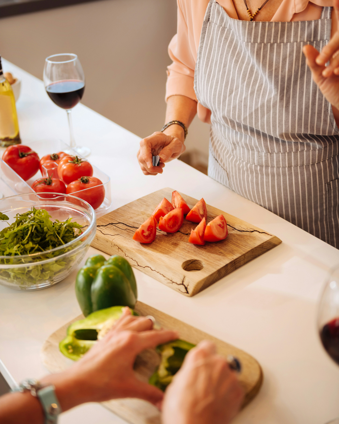 Hands slicing red peppers on a cutting board in a bright kitchen with fresh vegetables and wine nearby