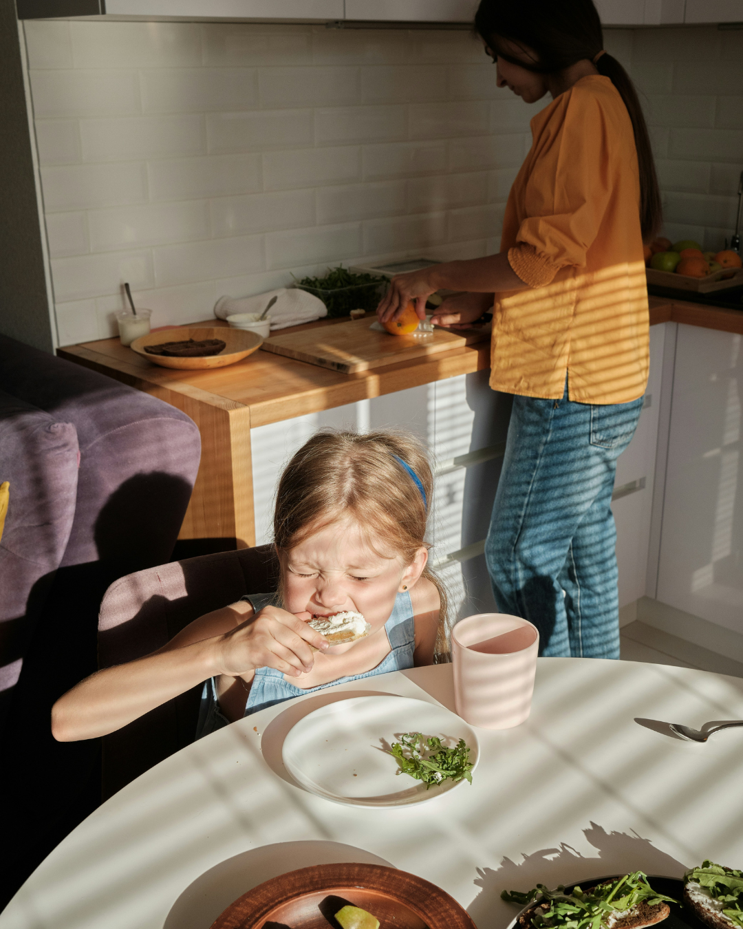 Child eating at a kitchen table while an adult prepares food at the counter in the background.