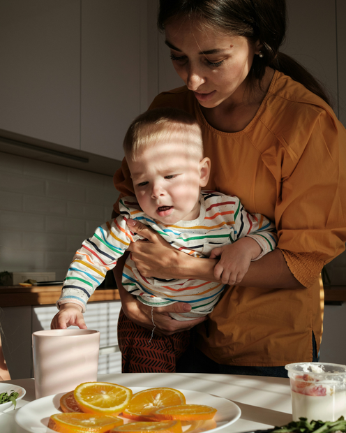 Woman guiding a toddler in a sunlit kitchen beside a table with oranges and yogurt cups