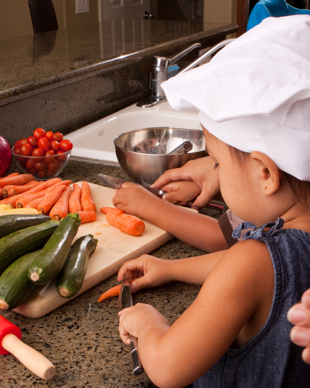 Child wearing a chef hat preparing vegetables at a kitchen counter with an adult nearby