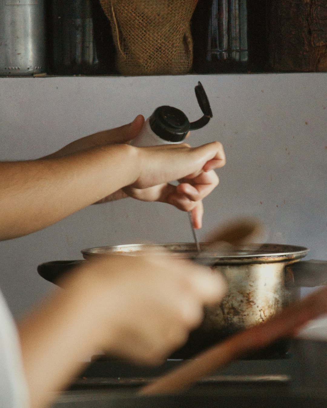 Hands stirring a pot on a stovetop in a kitchen, with a blurred pan in the foreground.