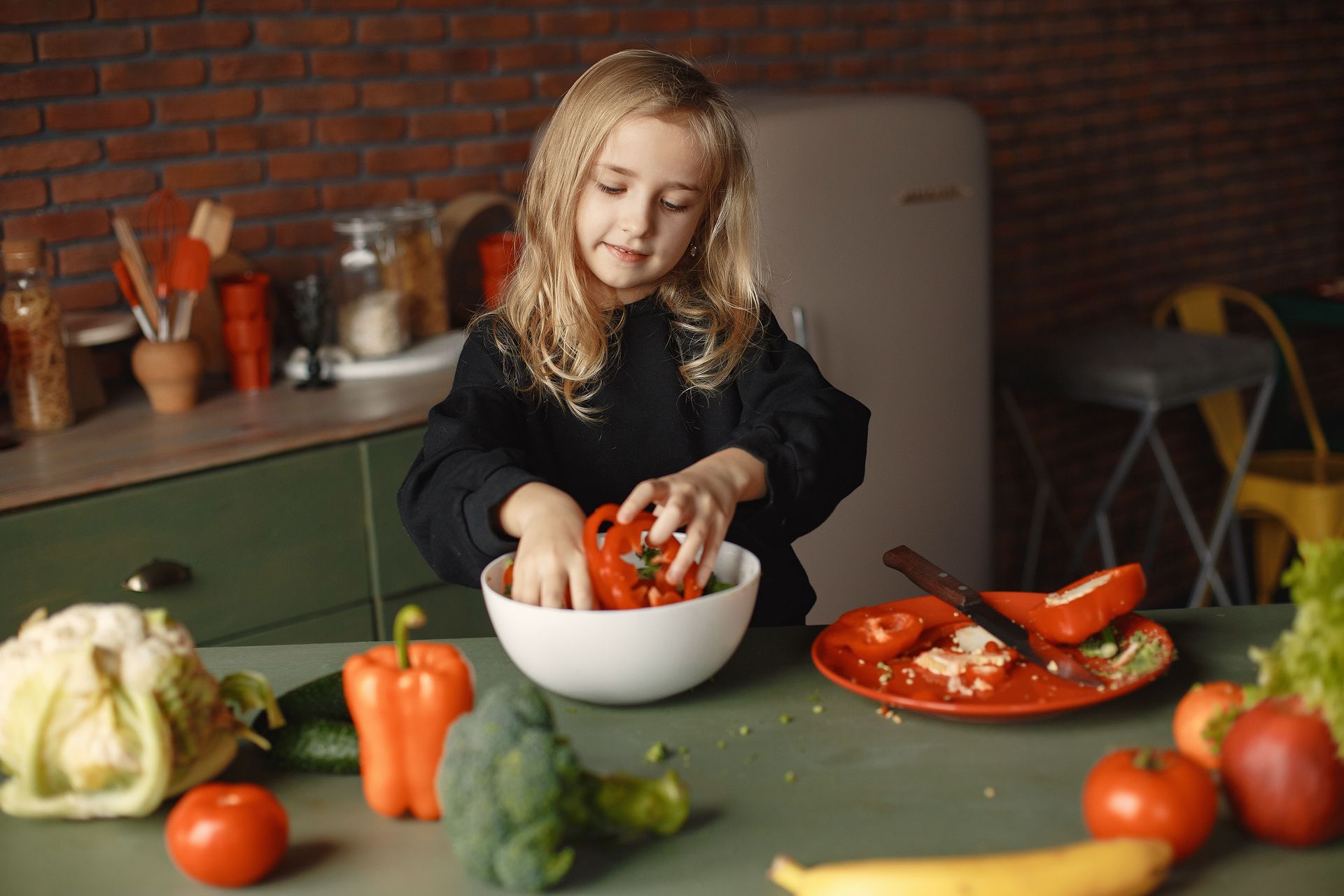 Child preparing vegetables at a kitchen counter with bowls of tomatoes and peppers, smiling softly.