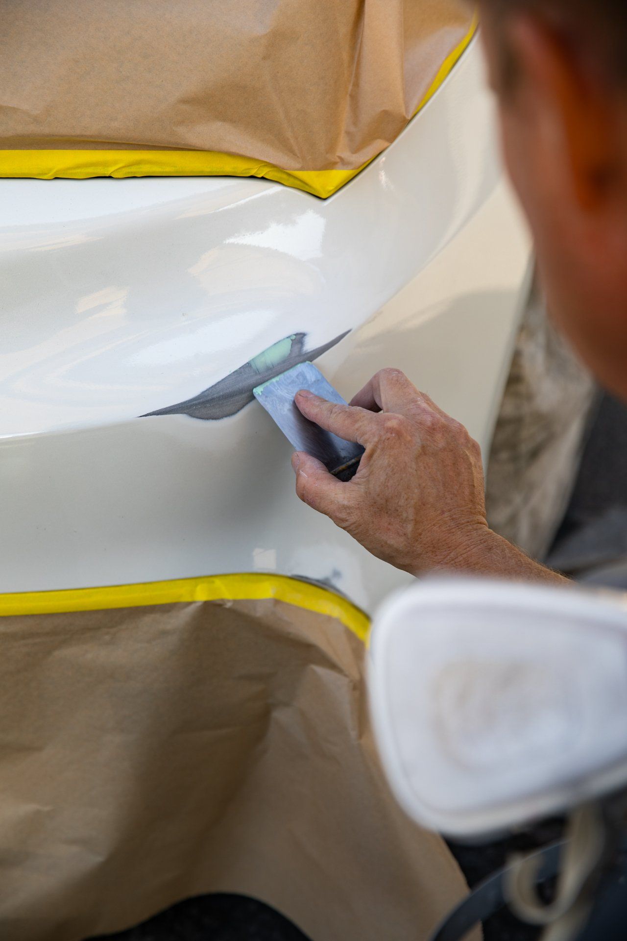 Person painting a white car panel with a small brush, masked with brown paper and yellow tape.
