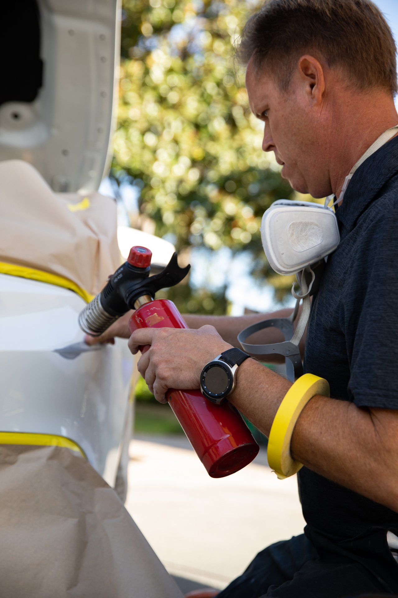 Person polishing a white car with a red cloth and spray bottle, wearing a respirator mask outdoors.