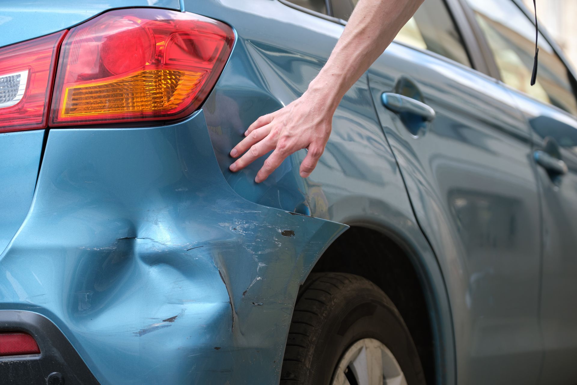 Hand touching a dented, light blue car. The rear panel is damaged and scratched.