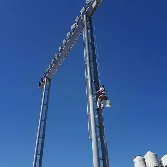 Un trabajador en un poste alto inspecciona las luces del estadio contra un cielo azul despejado.