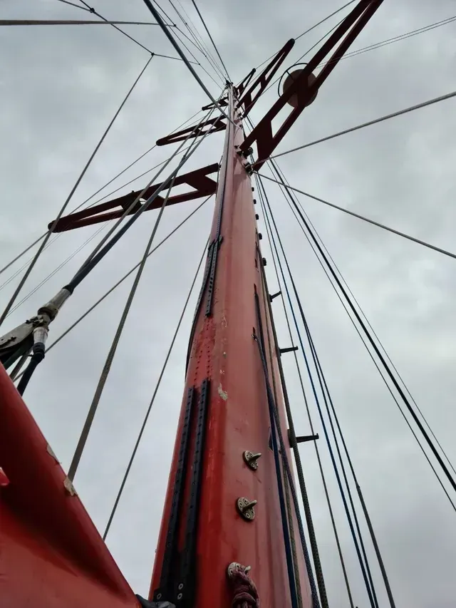 Mirando hacia arriba el mástil de un velero rojo con su aparejo contra un cielo nublado.