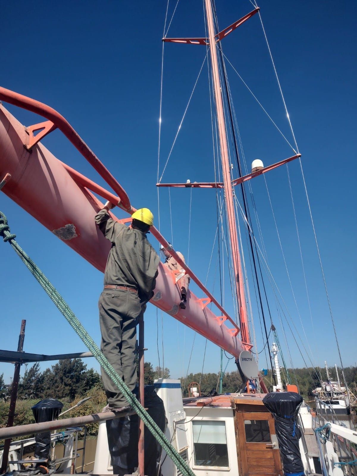 Un trabajador con casco amarillo, subido al mástil de un velero bajo un cielo azul, repara el aparejo en el muelle.