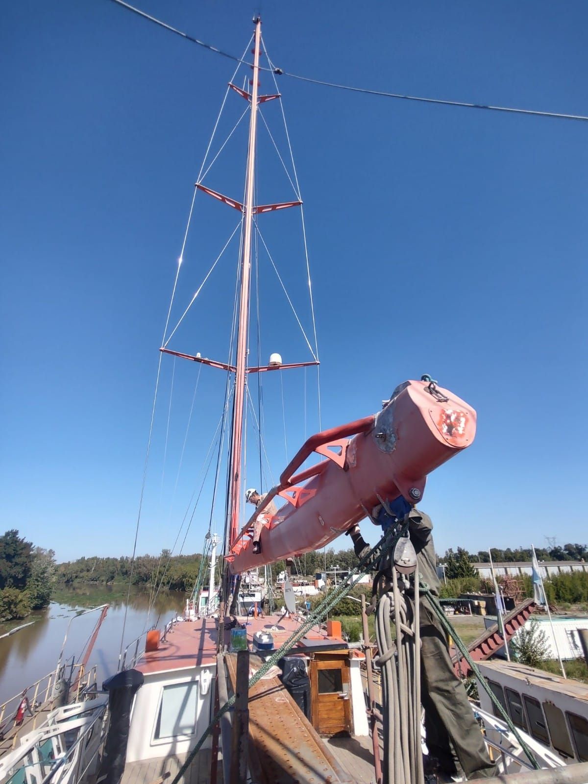 Mástil y aparejo de un velero rojo junto a un bote salvavidas rojo en un muelle bajo un cielo azul despejado.
