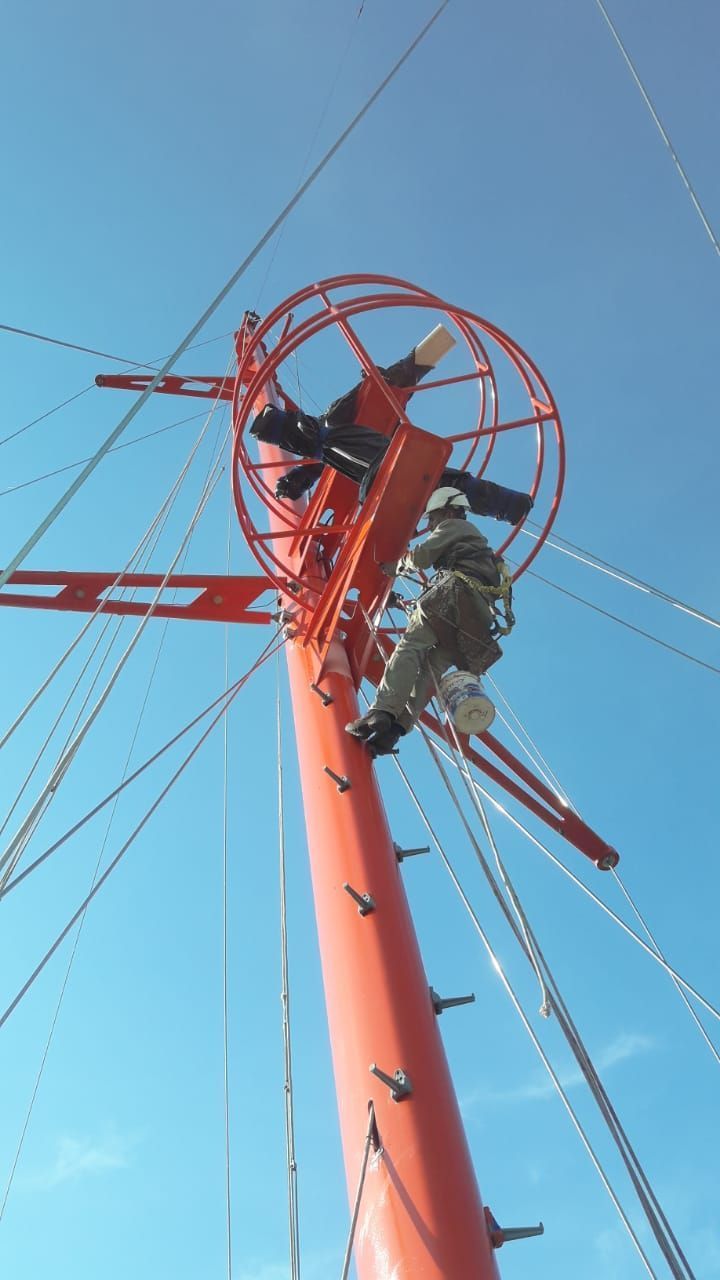 Un trabajador trepa a un alto mástil naranja con una plataforma circular contra un cielo azul.
