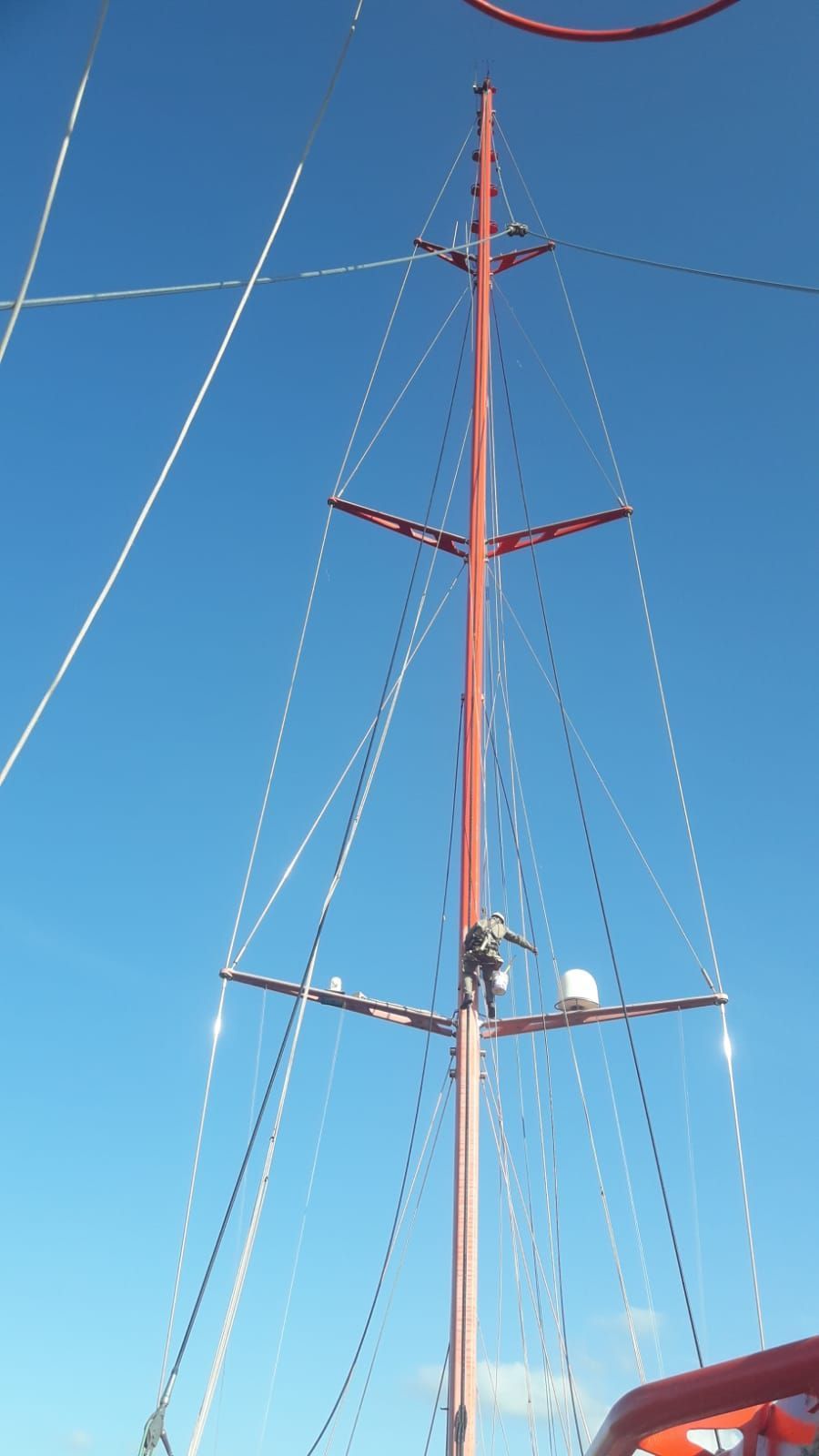 Alto mástil rojo de velero con su aparejo contra un cielo azul claro.