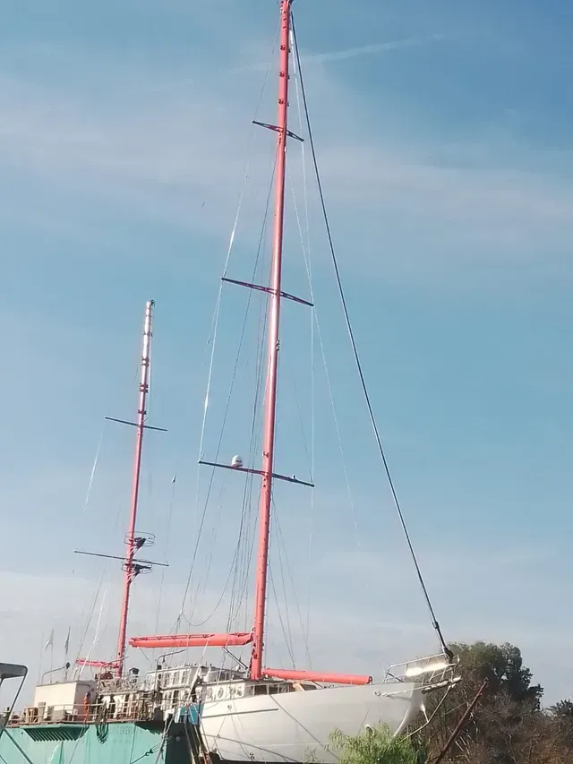 Dos mástiles de velero rojos con sus aparejos amarrados a la orilla bajo un cielo azul claro.