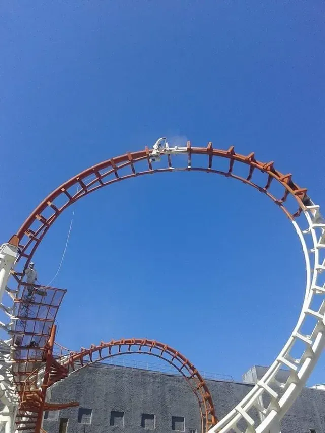 Vía de montaña rusa con bucles contra un cielo azul claro, con un edificio en primer plano.