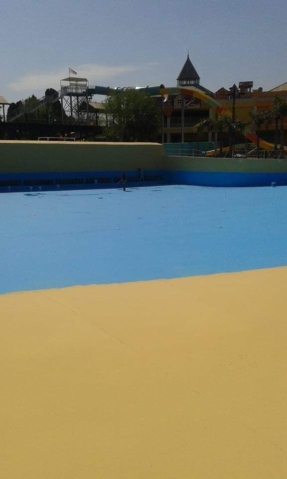 Piscina vacía de color azul y amarillo de un parque acuático, con toboganes y estructuras de juegos infantiles a lo lejos.
