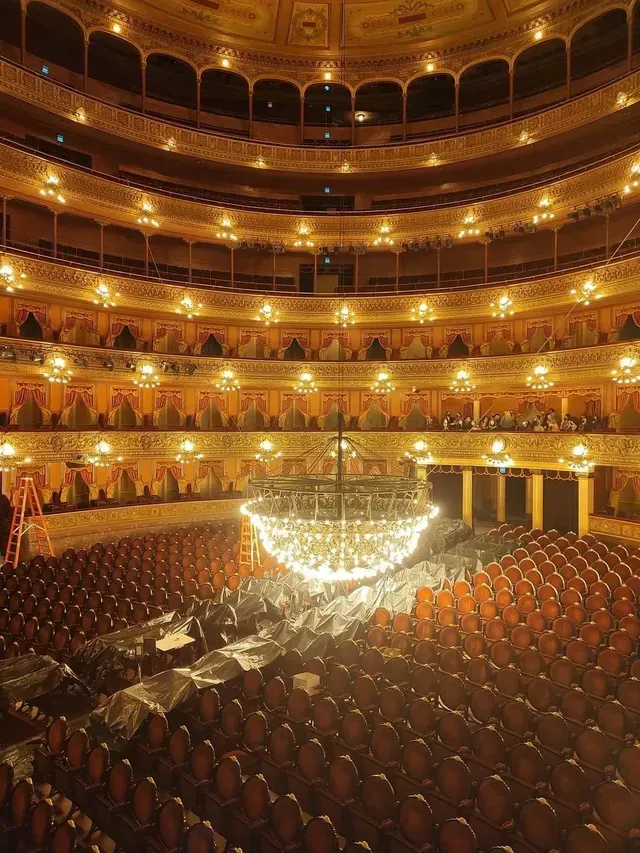Gran teatro ornamentado con butacas rojas y una lámpara de araña resplandeciente bajo balcones dorados.