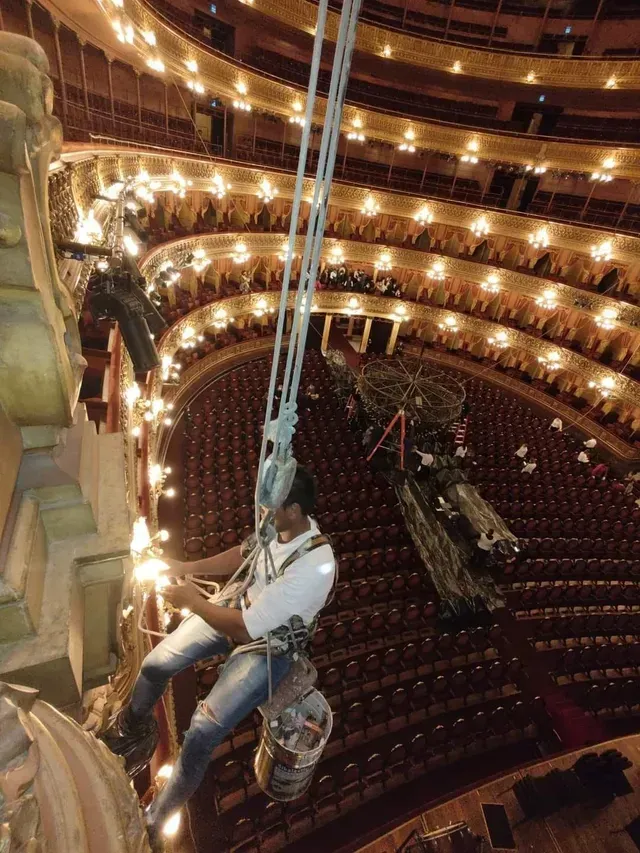 Una persona queda suspendida por cuerdas sobre el auditorio de un teatro con balcones ornamentados y luces cálidas.
