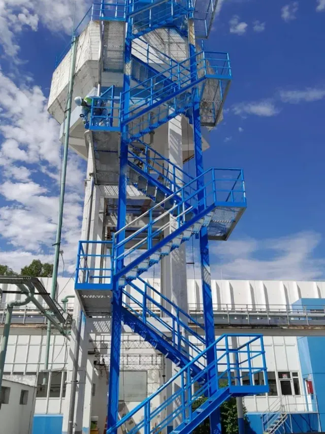 Escalera de caracol azul en una torre industrial junto a un edificio blanco bajo un cielo nublado.