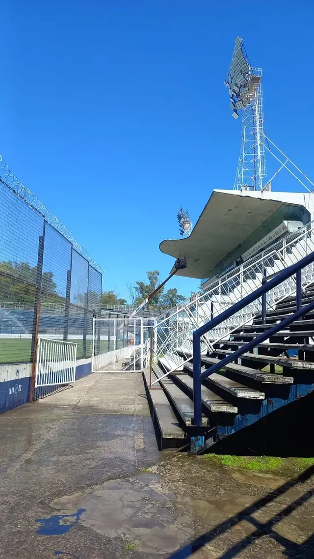 Gradas vacías de un estadio deportivo junto a una valla de tela metálica bajo un cielo azul brillante.