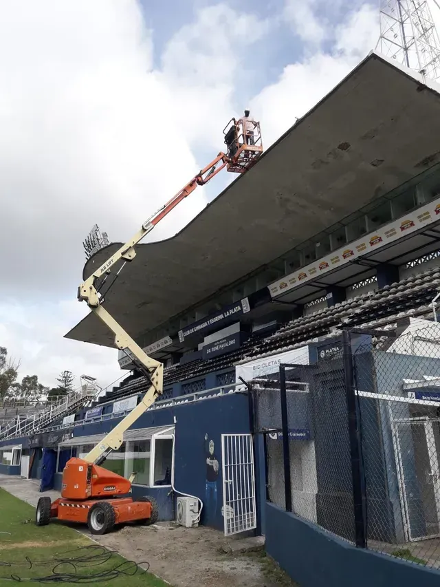 Un trabajador inspecciona el voladizo del techo de un estadio deportivo desde una plataforma elevadora.