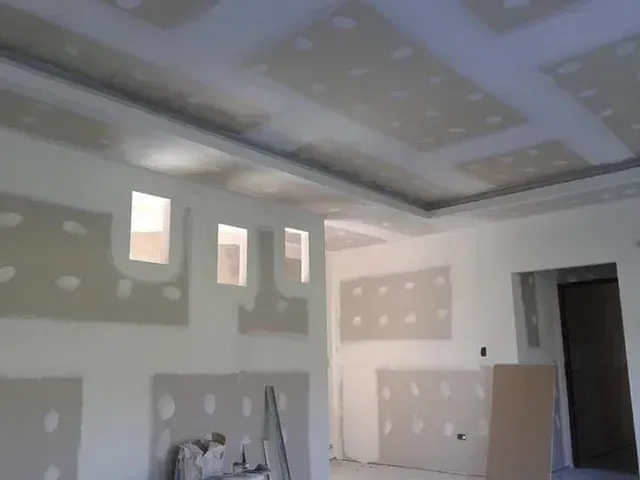 Habitación sin terminar con parches de paneles de yeso, ventanas y una escalera en proceso de renovación.