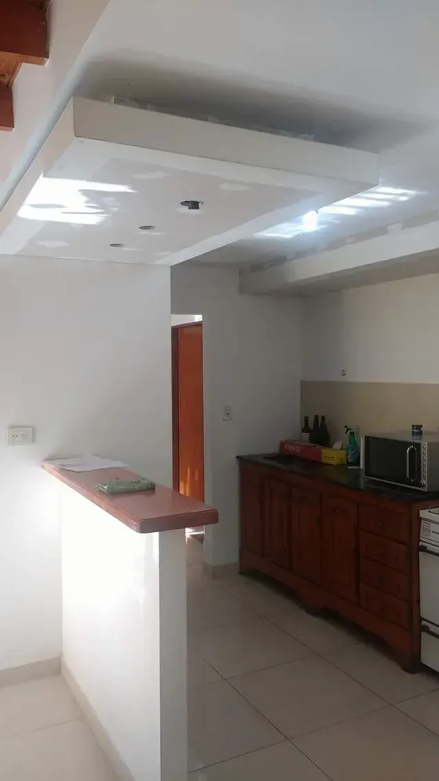 Cocina pequeña con paredes blancas, suelo de baldosas, armarios de madera y una barra pasaplatos bajo luces de techo.