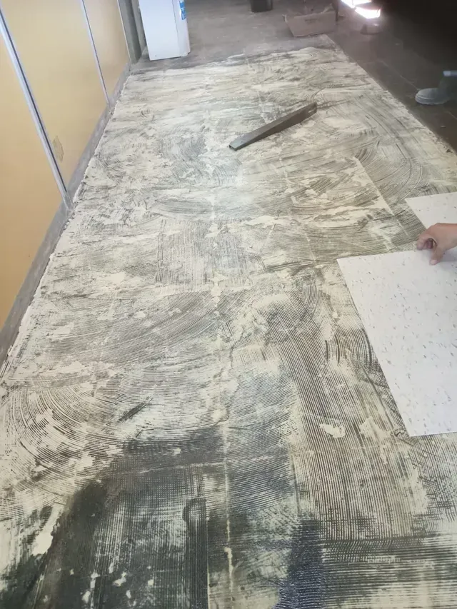 Suelo del pasillo dañado con restos de adhesivo antiguo y una mano sujetando un panel blanco a la derecha.