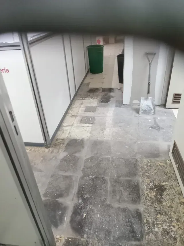 Pasillo interior estrecho con suelo de baldosas desgastadas, paredes blancas, un cubo de basura verde y herramientas.