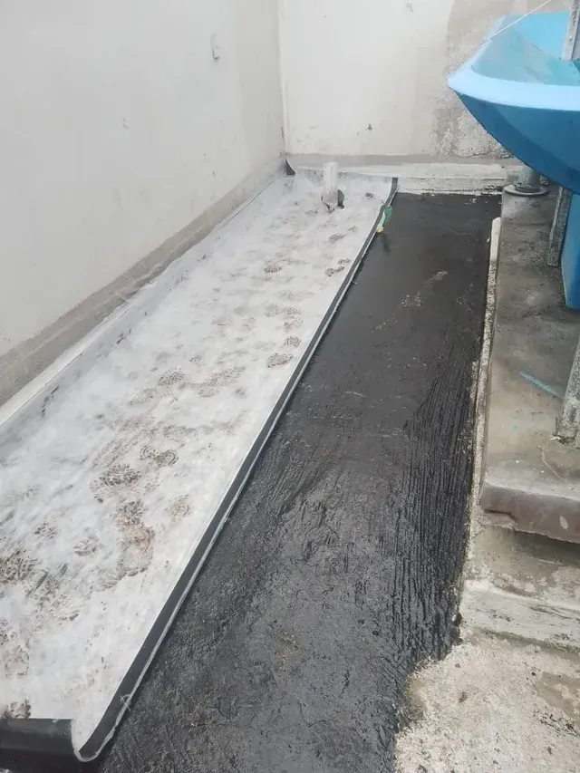 Pasarela estrecha al aire libre con hormigón fresco, pared blanca y un objeto azul en el borde derecho.