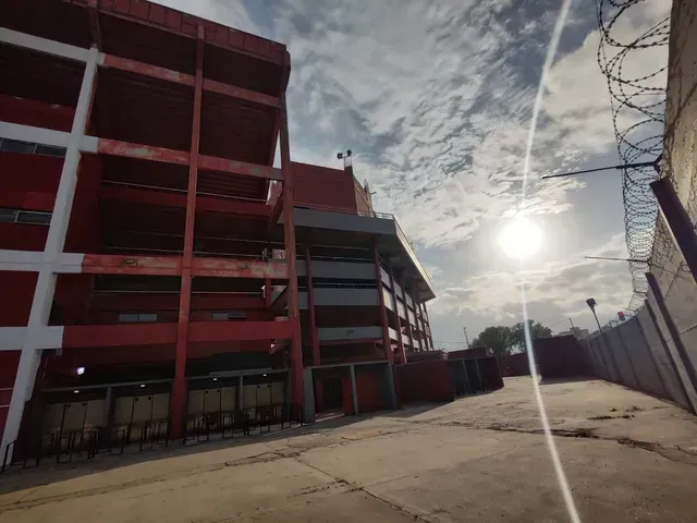 El estadio rojo se alza junto a una carretera bajo un sol radiante y un cielo nublado.