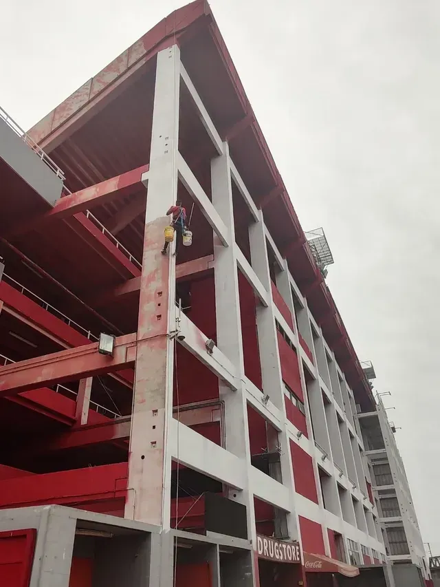 Estacionamiento de hormigón de varios pisos, de color rojo y blanco, en construcción, con un trabajador en un nivel superior.