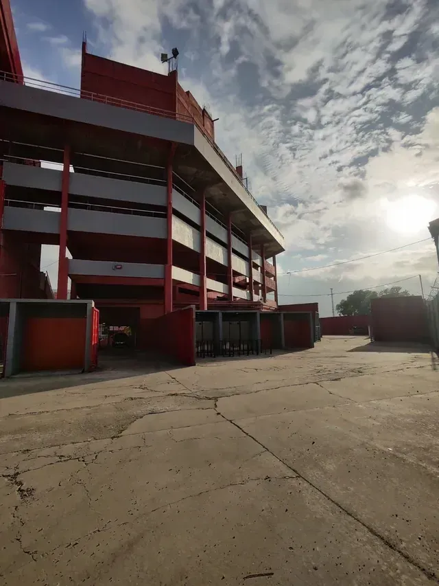 Edificio de varias plantas de color rojo y blanco junto a un solar vacío de hormigón bajo un cielo nublado.